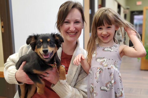 A woman and a girl smiling. The woman is holding a puppy in her arms